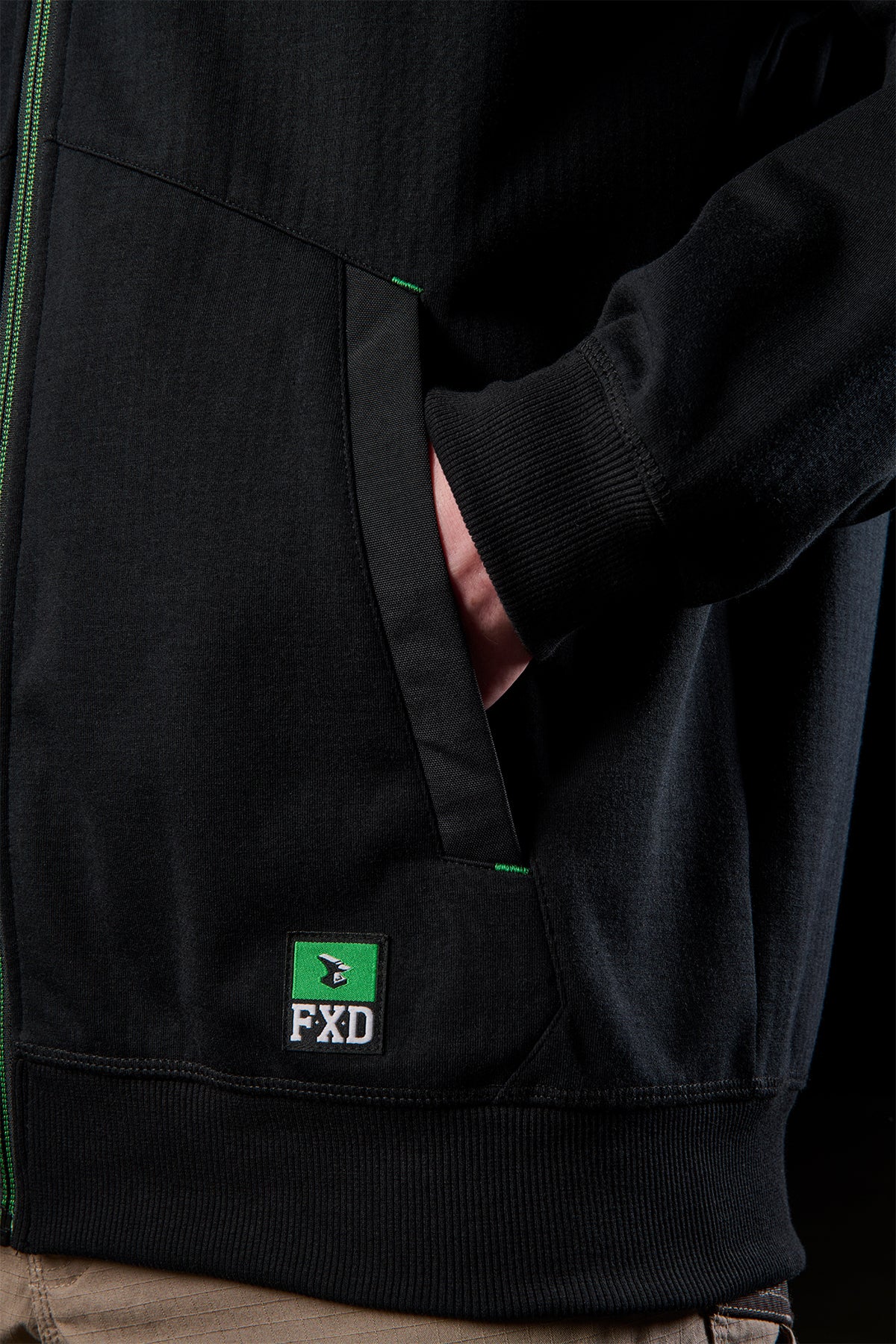 Close-up of a black WF-3 bonded fleece hoodie showing a zip pocket with green stitch details and FXD logo patch.
