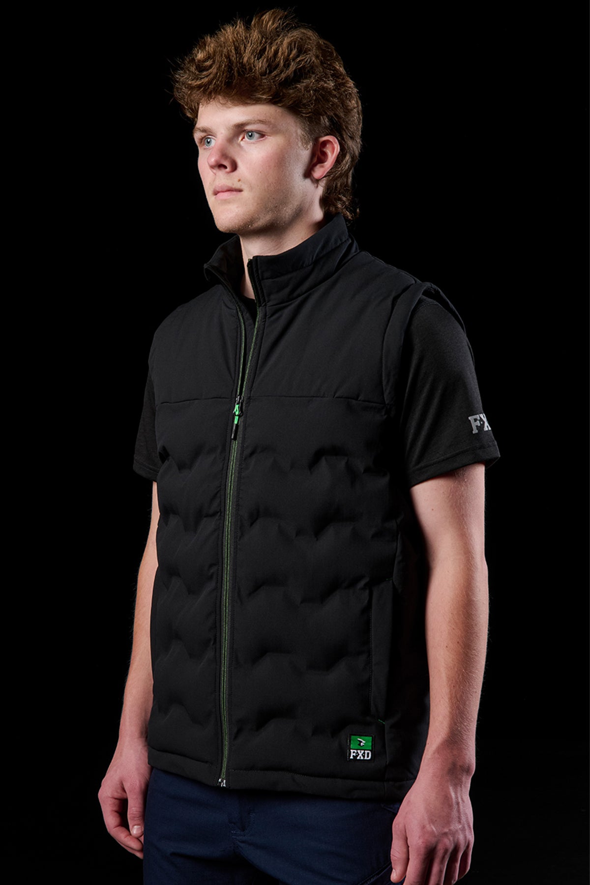 Side profile of a male model wearing FXD WO.6 insulated puffer vest with a green zipper over a black T shirt.