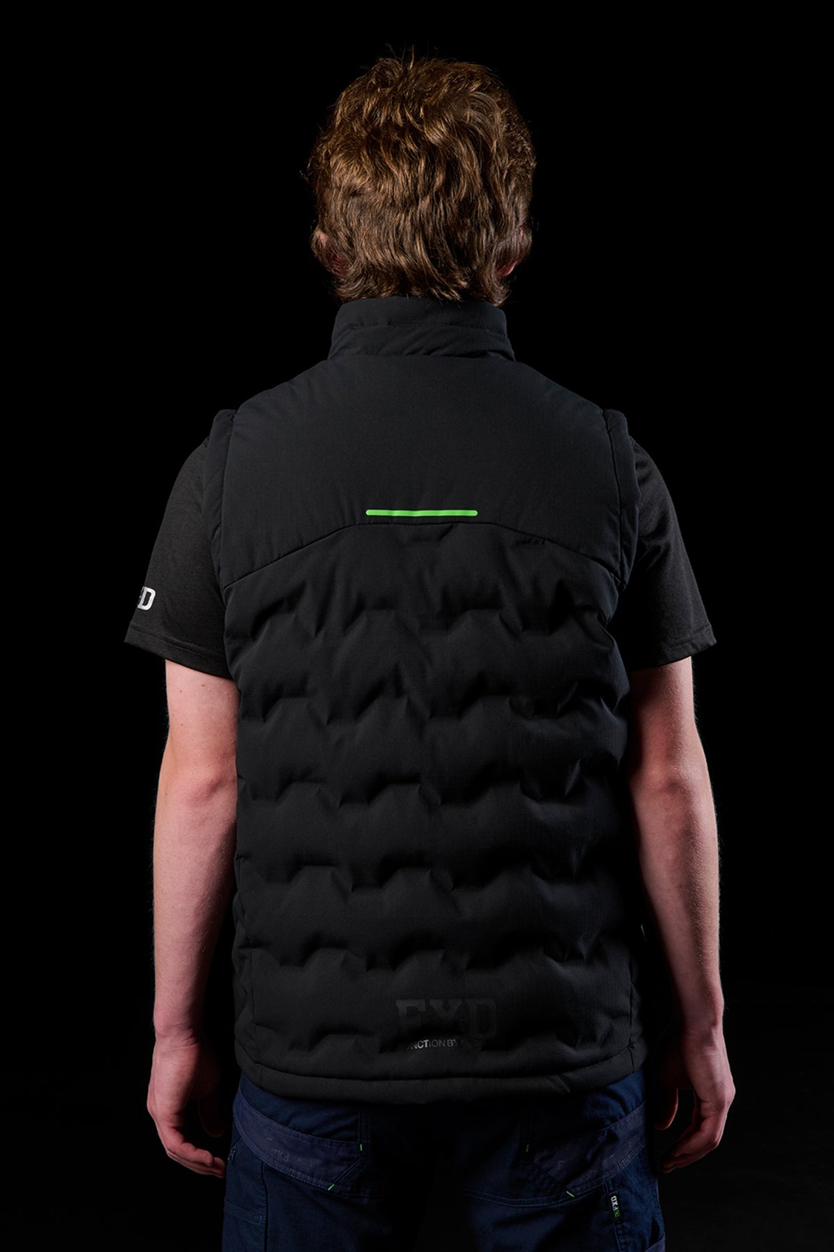 Back view of a model wearing a black FXD WO.6 insulated puffer vest with a lime green reflective strip across the upper back.