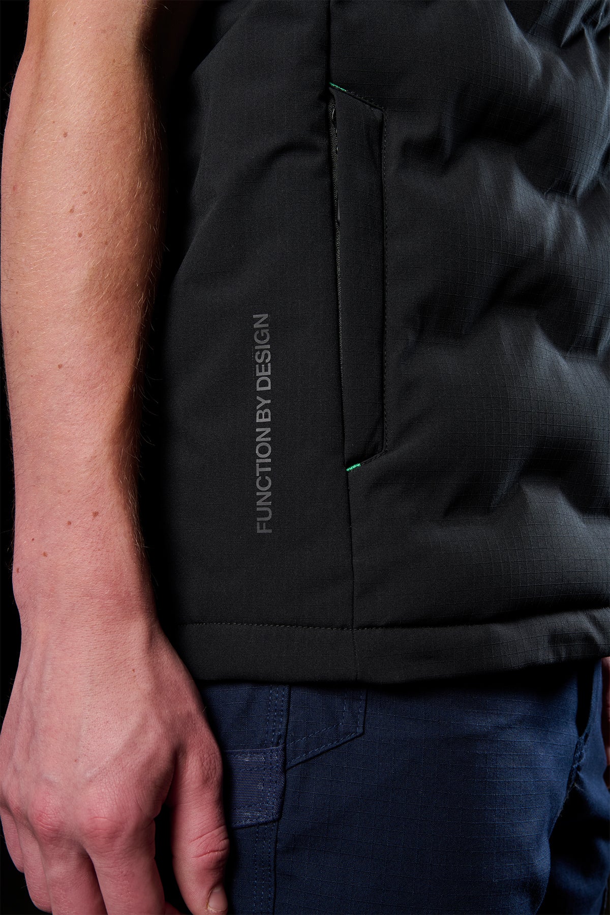 Close-up of black FXD WO.6 insulated puffer vest fabric showing a zippered side pocket and vertical Function by Design branding.